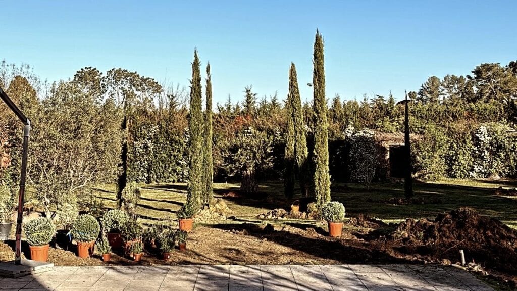 Aménagement paysager d'un jardin avec cyprès de Provence, plantes en pots et tas de terre sous un soleil radieux.