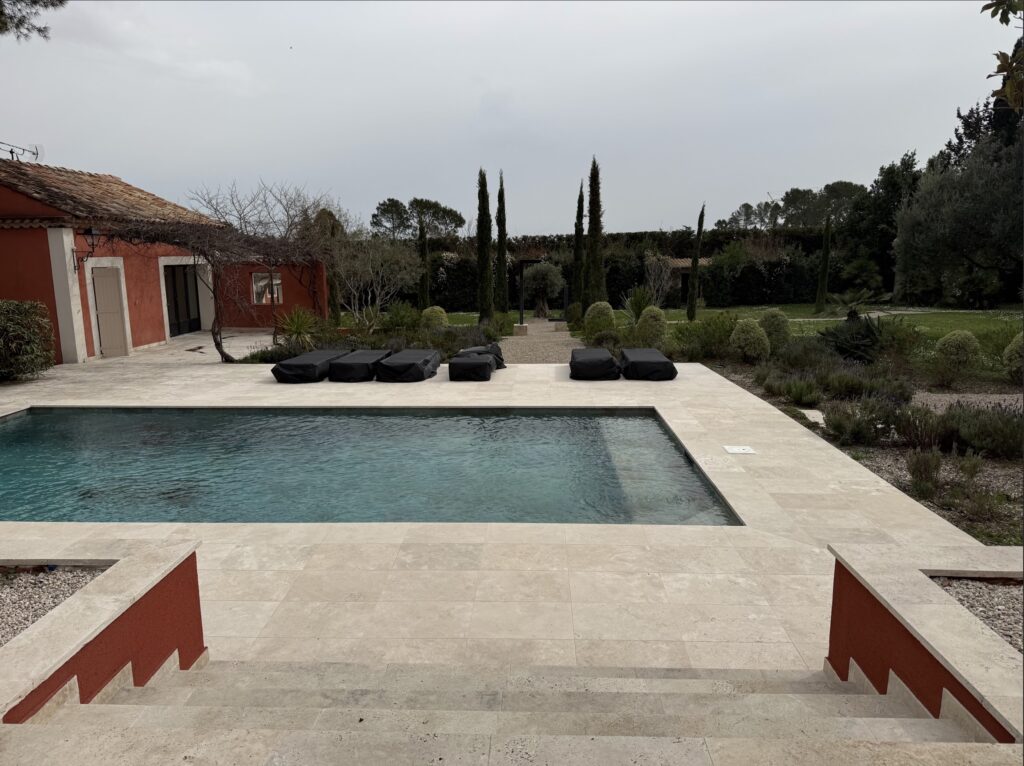 Piscine moderne avec terrasse en pierre, bains de soleil noirs et maison rouge provençale dans un jardin arboré.